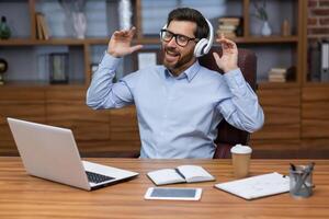 A young man businessman sits in the office at the table, works on a laptop in white headphones and listens to music, rests. sings along, dances. photo