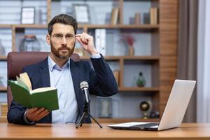 Online presentation of the book. Record a podcast. A young man writer, author, translator in a suit sits in the office at a table in front of a camera, a laptop and a microphone, holds a book. photo