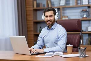 Portrait of a young male businessman working in the office at a laptop, sitting in white headphones and smiling at the camera. photo