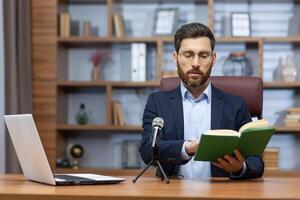 distancia aprendiendo. un joven masculino profesor se sienta en el oficina a un mesa en frente de un ordenador portátil y un micrófono y lee un en línea conferencia desde un libro a estudiantes, alumnos. foto