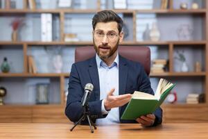 Senior mature pastor reading bible book online, man looking at camera sitting in home office and recording audio book online. photo