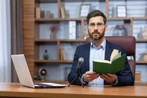 Portrait mature successful businessman teacher, man recording audio podcast reading book, boss glasses looking thoughtfully at camera working in home office with professional microphone and laptop photo