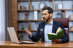 en línea presentación de el libro. un joven hombre escritor, autor en un traje se sienta en el oficina a un mesa en frente de un cámara, un ordenador portátil y un micrófono, sostiene un libro, lee a oyentes en un llamar. foto