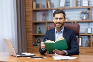 Joyful businessman in home office resting on break between work, mature man reading book, portrait of smiling boss holding book smiling and looking at camera. photo