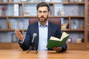 A man, a pastor of a church, a religious community of various denominations, conducts his sermon online. Sitting and speaking into a microphone in front of the camera, holding a holy book. photo
