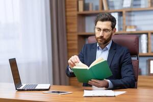 un joven hombre es sentado en el oficina en un traje a el mesa, participación y concentrando en leyendo un verde libro. descansando, estudiando, descanso. foto