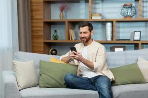 A mature man with a beard in a shirt at home is sitting on the sofa in the living room, using the phone, typing messages and browsing the Internet online. photo