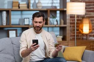 A young handsome man in a beige shirt is sitting on the sofa at home, using a phone and a credit card. photo