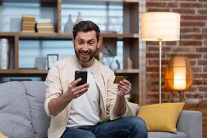 Satisfied and joyful buyer sitting on sofa at home, man using phone and bank credit card to buy online gifts in online store. photo