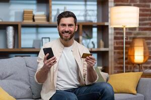 retrato de contento comprador en en línea almacenar, hombre sentado en sofá a hogar en vivo habitación sonriente contentamente y mirando a cámara participación teléfono y banco crédito tarjeta. foto