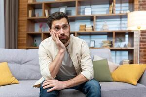 Overworked sad man sitting alone on sofa, mature businessman at home holding hands on head in depression in living room in casual clothes. photo