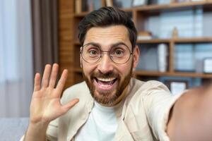 Young smiling man in glasses blogging during quarantine at home, talking on call, saying hello, holding online meeting, recording on camera. photo