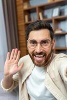 A young man in glasses talks at home on a call. He holds the phone in his hands, waves at the camera, smiles, says hello. Vertical photo. photo