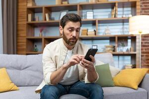 A man sitting on a couch at home, looking bored as he scrolls through dating apps on his phone. The casual setting and relaxed posture create a relatable image of modern dating culture photo