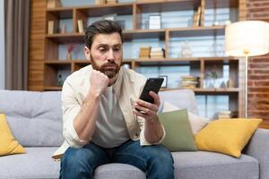 Sad and depressed man alone at home sitting on sofa, freelancer in casual clothes holding smartphone, unsatisfiedly reading bad news online from phone. photo