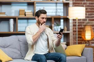 Sad and depressed man alone at home sitting on sofa, freelancer in casual clothes holding smartphone, unsatisfiedly reading bad news online from phone. photo