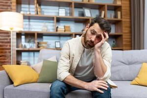 Overworked sad man sitting alone on sofa, mature businessman at home holding hands on head in depression in living room in casual clothes. photo