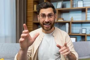 Mature teacher teaching online remotely, man with glasses and beard looking at web camera and smiling talking to colleagues and teaching, businessman at home sitting on sofa in living room. photo