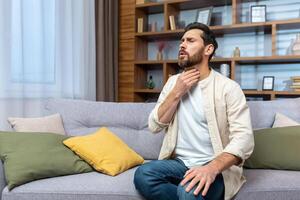 Sore throat. A young man sits at home on the sofa and holds his throat, feels a sore throat, burning sensation, discomfort. photo