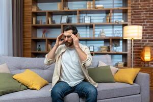 Headache of mature single man sitting on sofa alone at home and holding head with hands in living room. photo