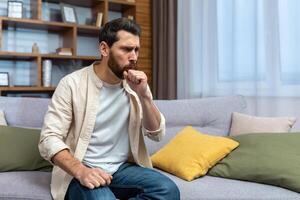 Sick mature man alone at home sitting on sofa coughing holding hands to chest in living room. photo