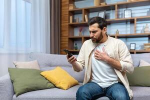 Mature man with beard alone at home reading bad news online on phone while sitting on sofa in living room at home. photo