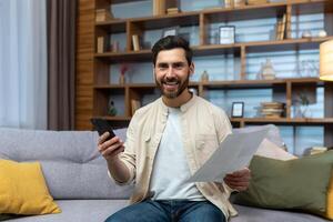 Portrait of a happy young man sitting at home on the couch. a smiling man looks at the camera, holds a phone and documents, papers in his hands. photo