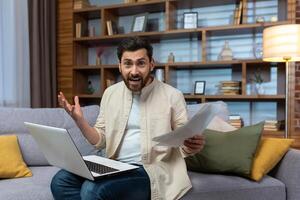 man alone at home sitting on sofa in living room, using laptop to calculate household budget, homework homework looking at camera and shouting tiredly. photo