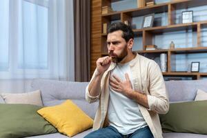 Sick mature man alone at home sitting on sofa coughing holding hands to chest in living room. photo