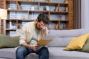 Upset man at home sitting on sofa reading letter with bad news notification in envelope opening, unhappy in living room and sad. photo