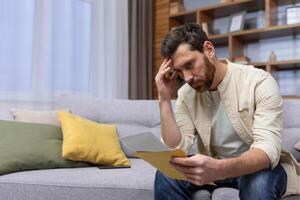 Upset man at home sitting on sofa reading letter with bad news notification in envelope opening, unhappy in living room and sad. photo