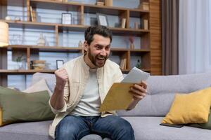 Joyful man at home holding envelope with notification message reading and smiling happy with news sitting on sofa inside living room. photo