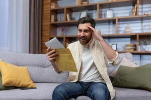 Joyful man at home holding envelope with notification message reading and smiling happy with news sitting on sofa inside living room. photo