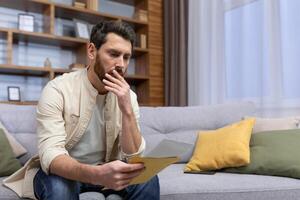 Shocked young man sitting on the sofa at home, holding a letter in his hands. Got bad news, divorce papers, financial debt, medical results. photo