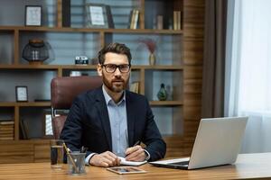 Portrait of serious mature businessman inside classic office at work, lawyer behind paper work looking focused at camera sitting at workplace using laptop and signing documents. photo