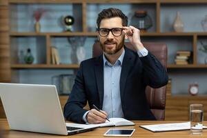 Portrait of successful smiling man in office, mature businessman looking at camera cheerfully, senior boss in glasses and beard working inside office with laptop and documents, signing contract. photo