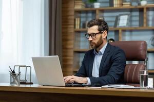 Successful mature businessman working inside office, man in business suit working at workplace with documents and laptop, boss behind paperwork attentive and focused. photo