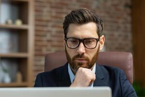 Close-up photo. Portrait of a young man in glasses and a suit of a lawyer, a legal defender who is serious sitting in the office, working on a laptop. Thoughtfully holding his chin and beard. photo