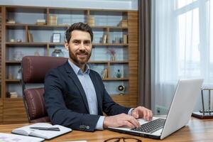 Portrait of mature successful boss at workplace, bearded man adult smiling and looking at camera, businessman in business suit inside office working with laptop, home office of lawyer financier. photo