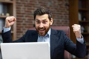 Successful businessman in home office celebrating victory and success, mature adult male boss satisfied with achievement results holding hands up triumph gesture financier happy using laptop at work photo