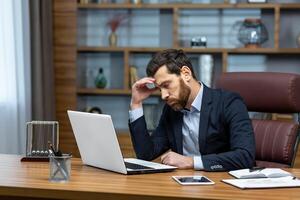 sad and depressed businessman in depression working inside modern office building, man in business suit with glasses and beard using laptop for work, boss disappointed with work result photo