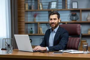 Portrait of mature successful boss at workplace, bearded man adult smiling and looking at camera, businessman in business suit inside office working with laptop, home office of lawyer financier. photo