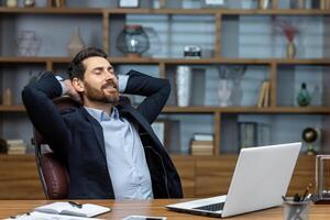 Completion work important project, mature man working inside modern office building using laptop, senior boss with beard and glasses putting hands behind head dreaming and visualizing future plans photo