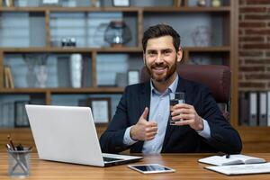 retrato de exitoso maduro empresario dentro oficina a lugar de trabajo, mayor experimentado hombre sonriente y mirando a cámara, utilizando computadora a trabajar, Bebiendo agua y demostración pulgares arriba alentador. foto