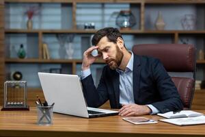 Serious thinking senior businessman working inside office using laptop, man in glasses solving problem, mature investor in business suit. photo