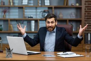 Angry boss yelling at camera, mature senior man working inside office, businessman in business suit with laptop nervous and angry. photo