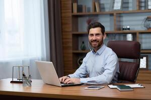 Portrait of successful businessman investor inside office, mature man smiling and looking at camera using laptop at work and wearing white headphones for call, photo