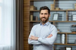 Portrait of successful businessman in office, man in shirt smiling and looking at camera, mature boss with beard with shaggy hands standing at workplace inside building. photo