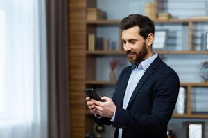Successful mature experienced man in business suit standing near window inside office, boss using phone, typing message and browsing online pages, businessman satisfied with work. photo
