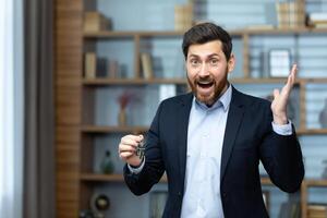 Portrait of a happy and successful realtor broker, the man looks joyfully at the camera and holds the keys to a new apartment in the house, a businessman in a business suit works inside the office. photo